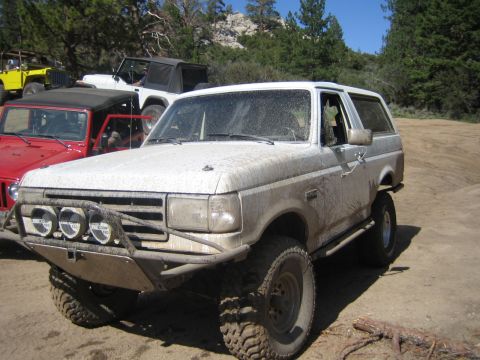 1990 Ford Bronco - mud-splattered two-tone Ford Bronco with front light bar sits on a dirt clearing beside red and yellow Jeeps among pines.