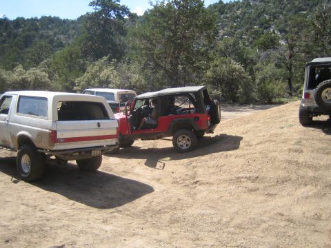 1990 Ford Bronco - red Jeep Wrangler with its doors removed sits on a dirt slope between a white Ford truck and a silver Jeep among trees.