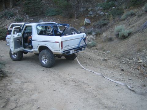 1990 Ford Bronco - white Ford Bronco with a blue roll cage, open door and trailing recovery strap is parked on a dirt mountain trail.