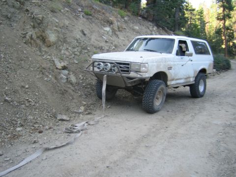 1990 Ford Bronco - mud-splattered white Ford Bronco with a front light bar and tube bumper sits on a dirt trail beside a rocky cut bank.