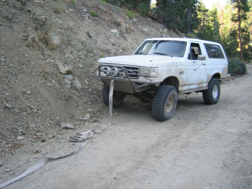 Mud-splattered white Ford Bronco with light bar on a dirt trail