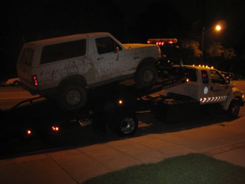 1990 Ford Bronco - muddy white Ford Bronco SUV strapped to a flatbed tow truck at night under a street light.