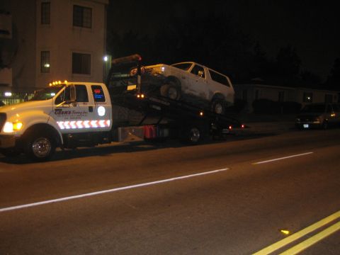 1990 Ford Bronco - white SUV loaded on a flatbed tow truck parked along a city street at night near apartment buildings.