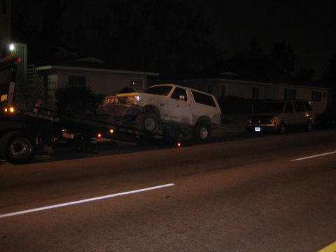 1990 Ford Bronco - mud-caked white SUV strapped onto a flatbed tow truck on a dark residential street at night.