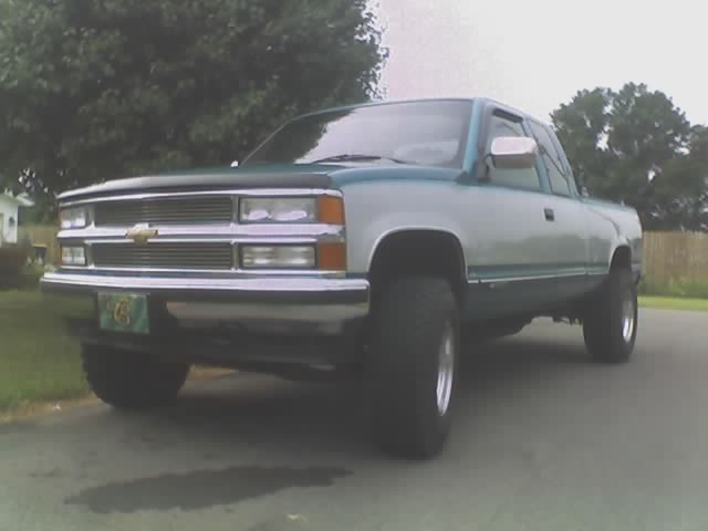 Teal and silver extended-cab Chevy pickup with billet grille, hood deflector, and chrome wheels parked on a street