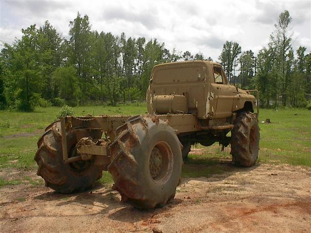 Mud-covered truck with oversized tires, missing its bed, viewed from the rear three-quarter angle