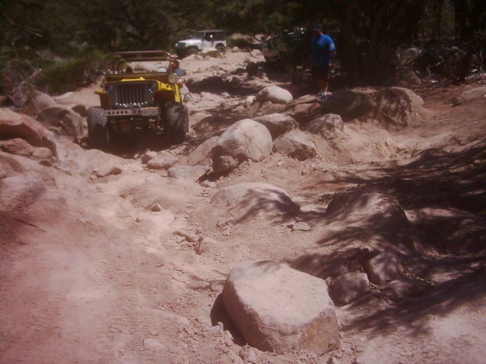 Yellow jeep and white jeep driving on a rocky mountain trail. A person stands on the side