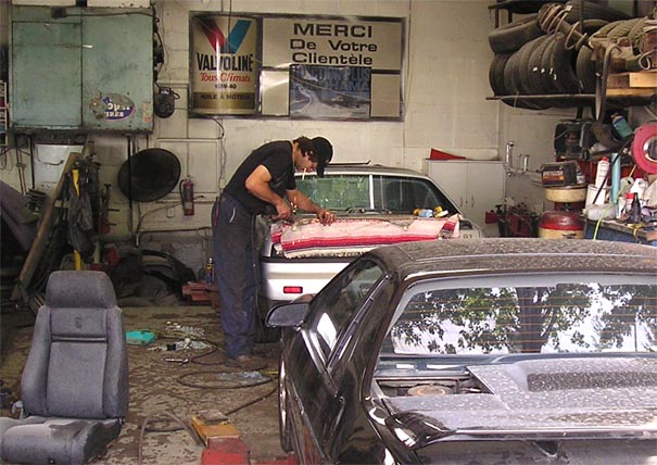 Two cars, one black and one silver, in a cluttered garage with a mechanic working on the silver car