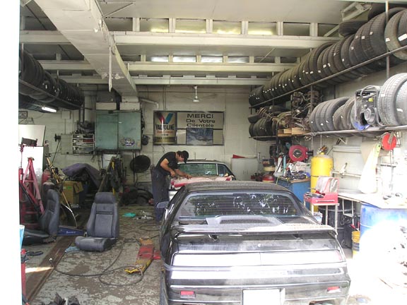 Black Pontiac Fiero in a garage with tires stacked on shelves and a mechanic working on another car