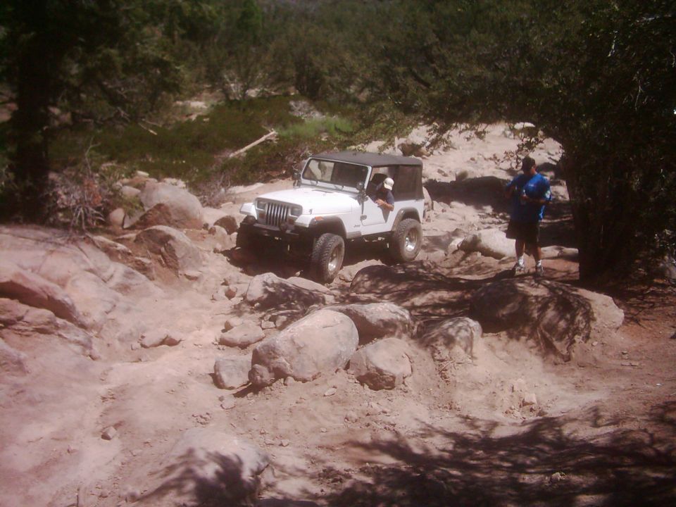 White Jeep Wrangler navigating a rough, rocky off-road trail with a person standing nearby