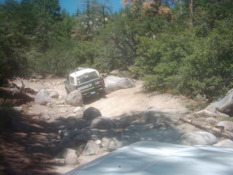 1978 Ford Bronco - A white SUV navigates a challenging rocky path through a wooded area, with another vehicle following behind.