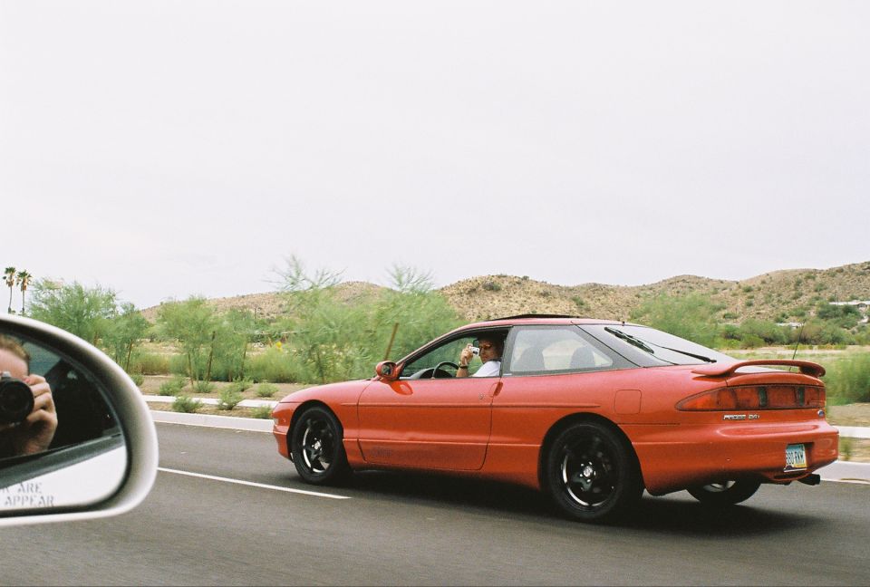 Red Ford Probe coupe with black wheels on a highway, driver photographing back; photographer visible in side mirror.