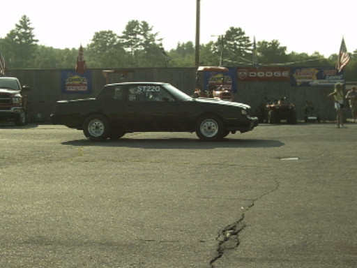 Black coupe with ST220 on rear window parked at a drag strip with sponsor banners behind