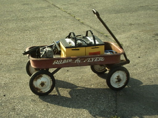 Red Radio Flyer wagon holding two batteries and a small engine on pavement