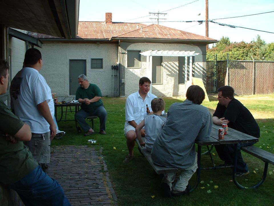 Several guys hang out in a backyard, some sitting at picnic tables, others standing. A house and fence are in the background