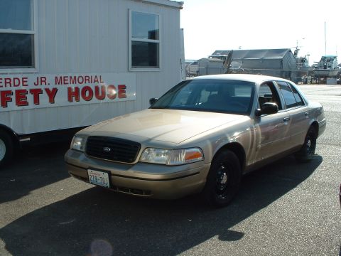 1999 Ford Crown Victoria - A tan 1999 Ford Crown Victoria sedan is parked in front of a trailer that reads 'VERDE JR. MEMORIAL SAFETY HOUSE'.