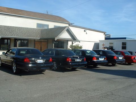 1999 Ford Crown Victoria - Several black sedans are parked in a lot outside a building on a sunny day.