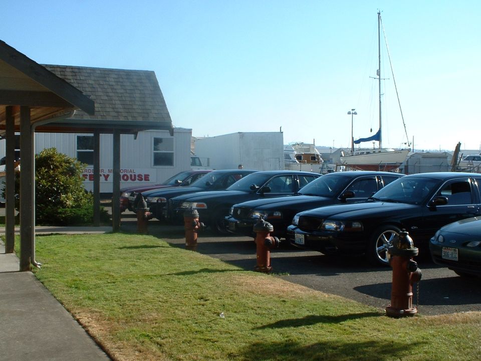 Row of black sedans parked on asphalt, with a building and boats in the background