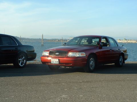 1999 Ford Crown Victoria - Two Ford Crown Victorias are parked near a body of water with a clear sky.