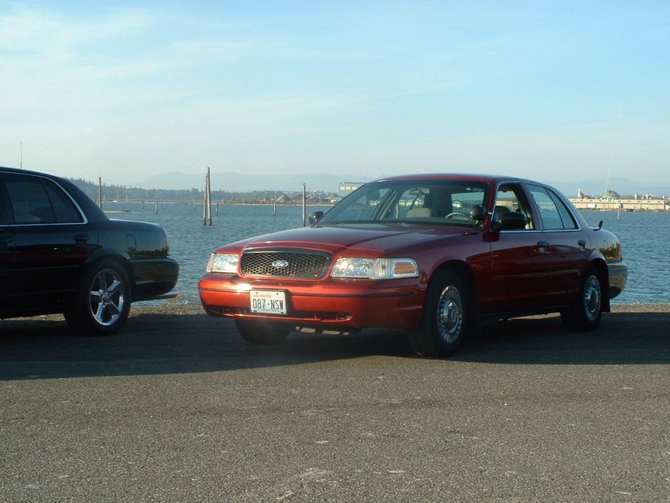 Red 1998 Ford Crown Victoria parked next to a black Crown Victoria, waterfront background