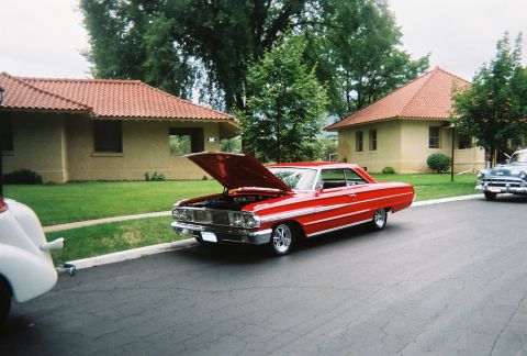 1972 Ford Ranchero - A bright red 1964 Ford Galaxie 500 is parked with its hood up, alongside another vehicle in front of two houses.