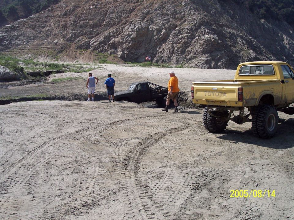 Yellow Toyota pickup truck near a black truck stuck in mud, with people standing around