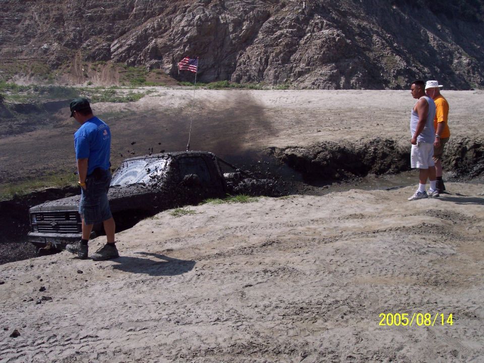 Truck stuck in mud pit, front grille visible, American flag on antenna