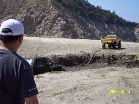 1985 Ford Bronco - Two off-road vehicles are stuck in a muddy pit, with a yellow truck attempting to pull one out.
