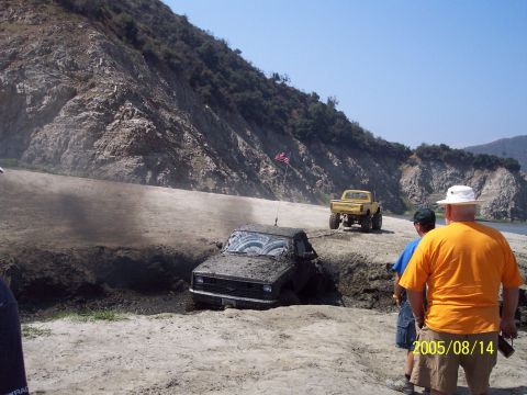 1985 Ford Bronco - Two trucks off-roading, one stuck deep in mud near a steep embankment.