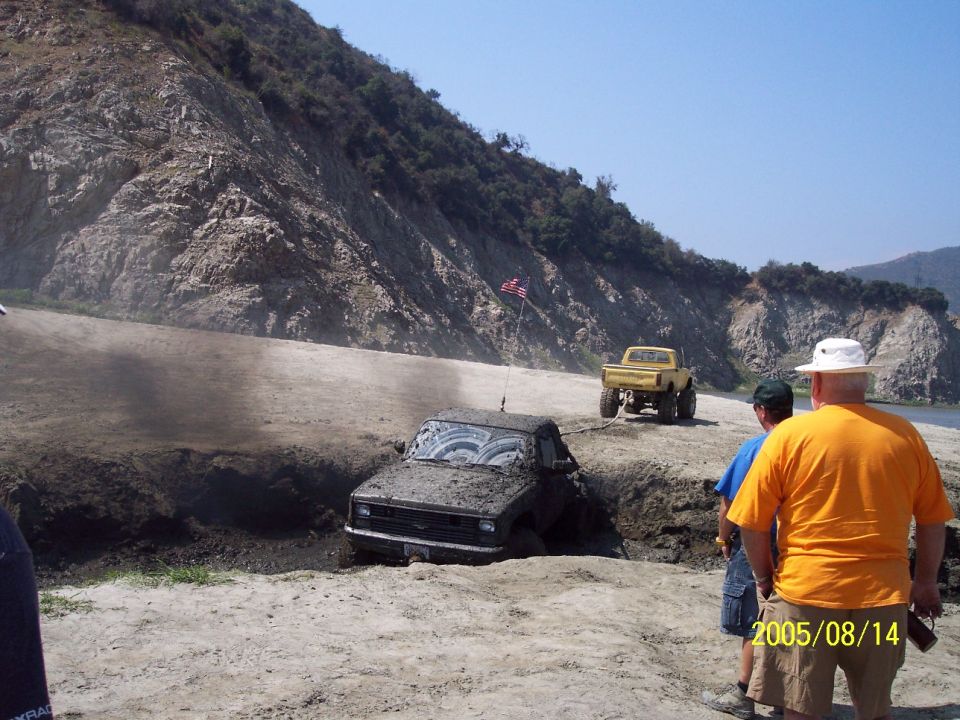 Mud-covered truck stuck in a ditch, yellow truck nearby, people watching, American flag on a pole