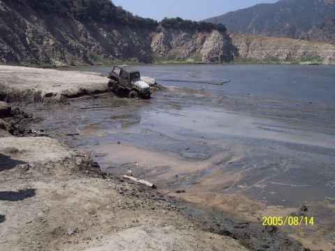 1985 Ford Bronco - A vehicle is stuck in the mud near the edge of a lake, with a steep, rocky hillside behind it.