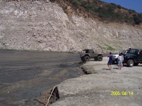 1985 Ford Bronco - Several off-road vehicles are parked on a dirt bank near a muddy area, with a rocky hillside providing a backdrop.