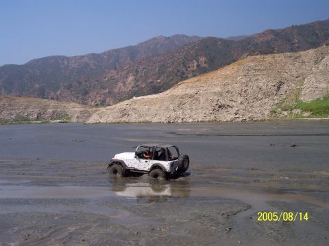1985 Ford Bronco - A white Jeep Wrangler is stuck in a muddy area with a mountainous landscape behind it.