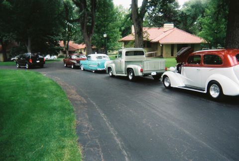 1972 Ford Ranchero - A collection of vintage vehicles are parked in a line on a paved driveway next to a grassy lawn and a building.