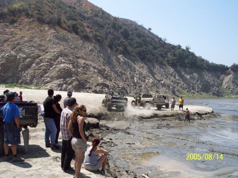 1985 Ford Bronco - A group of people watch as several trucks navigate a muddy off-road course on a sunny day.