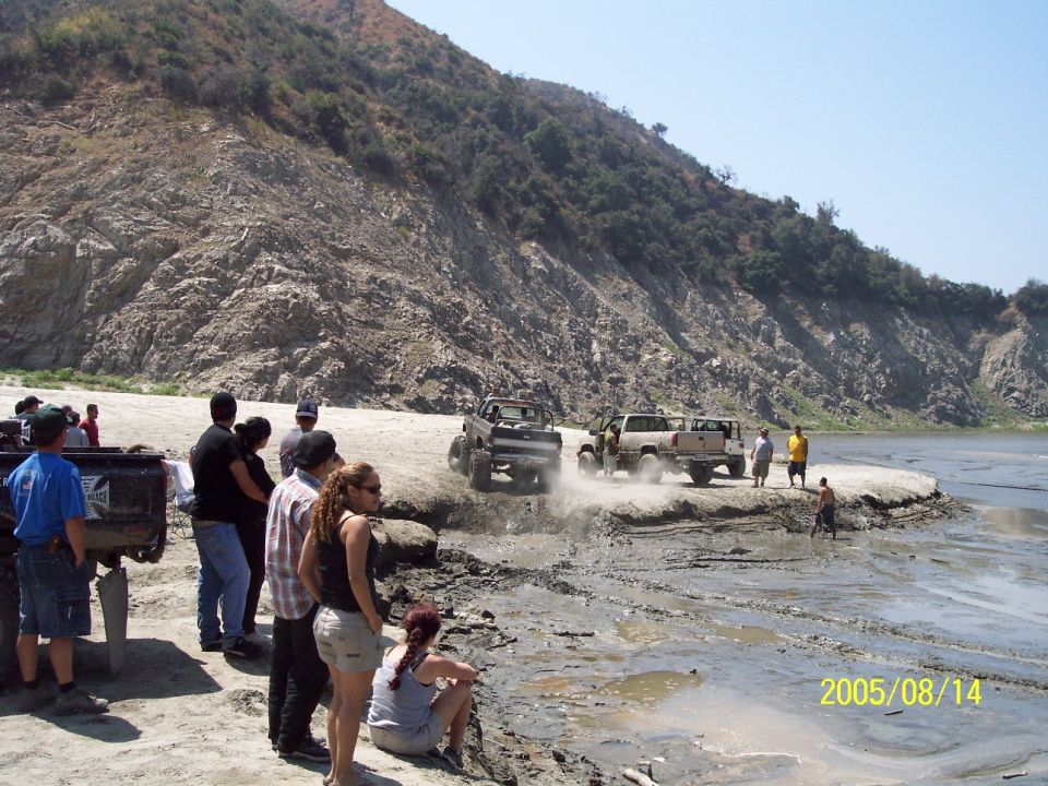Several off-road trucks driving through mud near a rocky hill and a body of water. People are watching