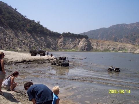 1985 Ford Bronco - Several off-road vehicles are stuck in the mud near a lake, with onlookers observing the scene.