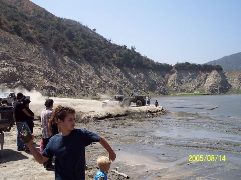 1985 Ford Bronco - A group of people observe off-road vehicles maneuvering in a dusty area next to a body of water on a sunny day.
