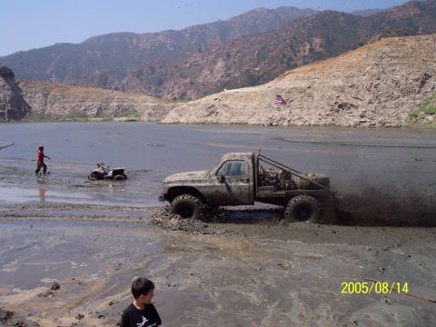 1985 Ford Bronco - Pickup truck and ATV navigate muddy terrain near a body of water with hills in the background.