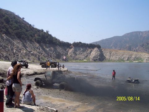 1985 Ford Bronco - Several off-road vehicles are seen kicking up mud near a body of water with spectators on the shore.