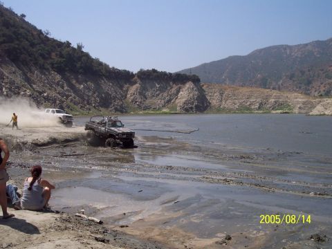 1985 Ford Bronco - Off-road vehicles navigate a muddy terrain next to a lake under a clear sky.