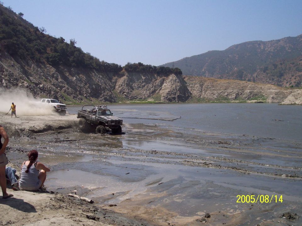 Two off-road trucks driving through mud near a body of water with a rocky hillside