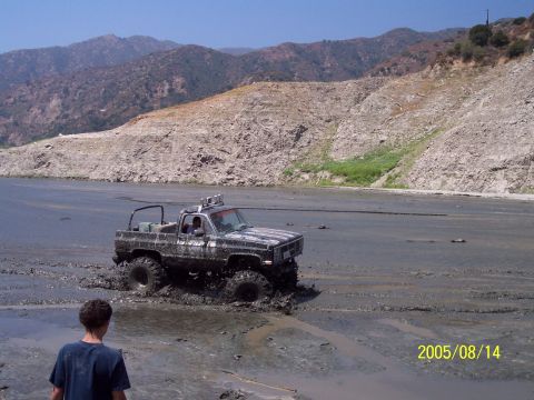 1985 Ford Bronco - A lifted pickup truck navigates through a muddy terrain, with a person watching in the foreground and mountains in the background.