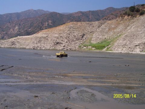 1985 Ford Bronco - A yellow pickup truck navigates a muddy terrain, with a mountainous landscape visible behind it.