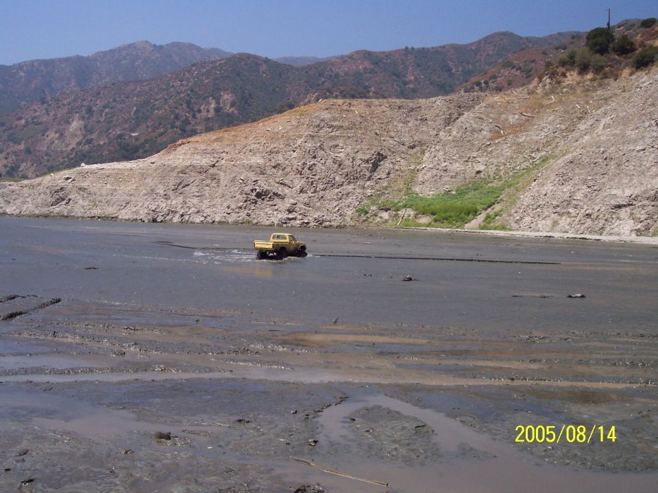 Yellow pickup truck driving through a muddy, dry lakebed with mountains in the background. Date stamp reads 2005/08/14