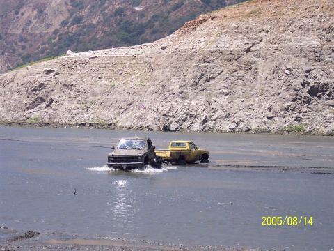 1985 Ford Bronco - Two trucks navigate through a body of water on a sunny day, with a rocky embankment behind them.