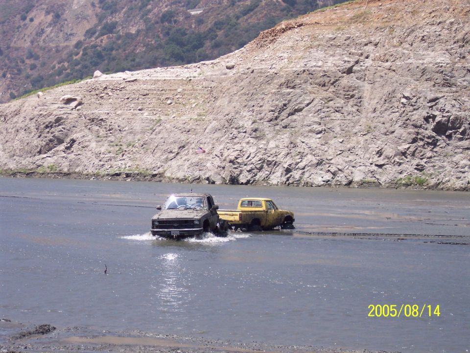 Two trucks driving through shallow water with a rocky hillside in the background