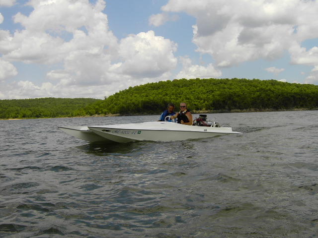 White catamaran boat with two people on a lake, trees in background