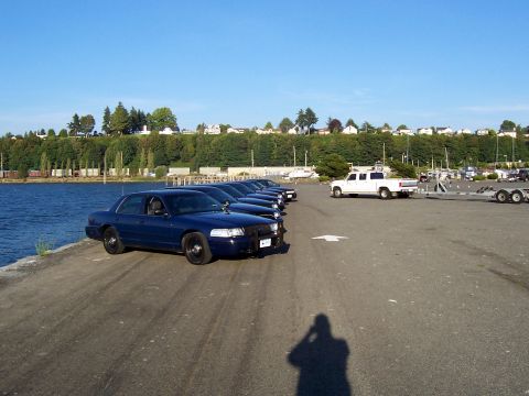 2003 Ford Crown Victoria - A line of dark blue sedans, likely police vehicles, are parked along a waterfront area on a sunny day.