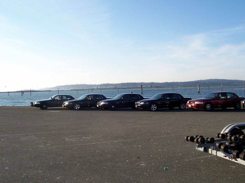 Side view of five Ford Crown Victorias, one red, four black, parked by the water